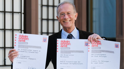 Der Chef der Staatskanzlei, Axel Wintermeyer, hält die drei Förderbescheide in die Kamera und lächelt.