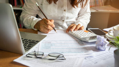 Eine Frau sitzt an einem Schreibtisch, vor ihr ein Laptop, Unterlagen und ein Taschenrechner.