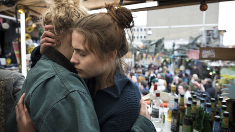 Zwei Frauen umarmen sich. Im Hintergrund sieht man Flaschen und mehrere Menschen