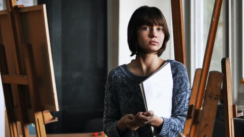 Eine Künstlerin in ihrem Atelier, sie hält einen Zeichenblock in der Hand und schaut ernst