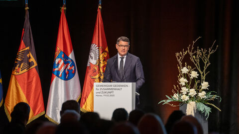 Ministerpräsident Boris Rhein steht an einem Rednerpult und spricht. Im Hintergrund stehen die Flagge von Deutschland, Hessen und Hanau.